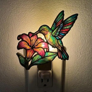 May include: A plug-in night light featuring a stained glass style design of a multi-coloured hummingbird hovering over a large, vibrant orange and pink lily flower with green leaves. The light emits a warm glow from behind the decorative panel.