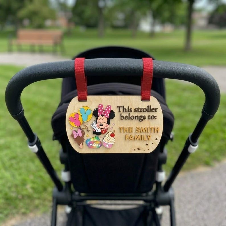 May include: A light-colored wooden stroller sign with red fabric straps, featuring a Minnie Mouse graphic, colorful balloons, a cupcake, and an ice cream treat. The text reads: This stroller belongs to: THE SMITH FAMILY, with small star accents.
