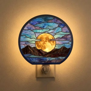 May include: A circular stained glass night light featuring a glowing yellow full moon set against a dark mountain range. The sky is composed of blue and purple glass segments with cloud-like patterns, all encased in a dark metal frame.