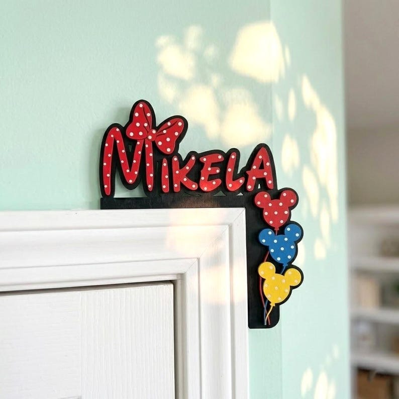 K&ouml;nnte beinhalten: Ein schwarzes T&uuml;r-Eckschild aus Holz mit dem Namen Mikela in roten Buchstaben mit wei&szlig;en Punkten und einer passenden roten Schleife. Unter dem Namen sind drei mausf&ouml;rmige Luftballons in Rot, Blau und Gelb mit wei&szlig;en Punkten angebracht.