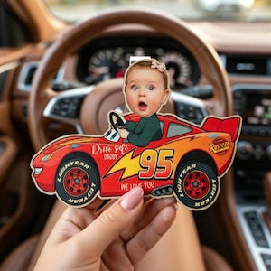 Op de afbeelding: Een houten auto-vormig ornament met een babygezicht als bestuurder. De auto is rood en oranje met het nummer 95 en de tekst "Drive safe Daddy" en "We love you". De wielen zeggen "Lightyear".