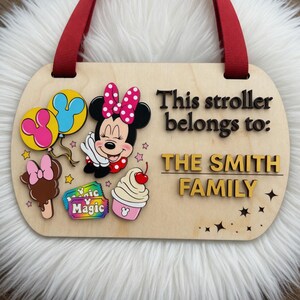 May include: A light-colored wooden oval stroller sign with a red ribbon hanger. It features a cartoon mouse with a pink polka-dot bow, two balloons, a chocolate treat, two tickets labeled Magic, and a sundae. Text reads: This stroller belongs to: THE SMITH FAMILY.