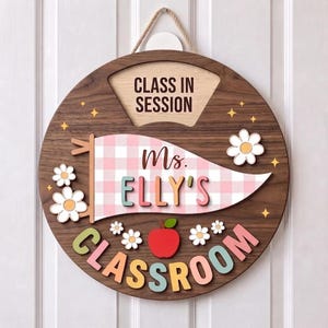 May include: A round wooden classroom door sign featuring the text CLASS IN SESSION at the top. A central pink and white checkered pennant displays the name Ms. ELLY'S above the word CLASSROOM, accented with white daisy cutouts and a red apple.