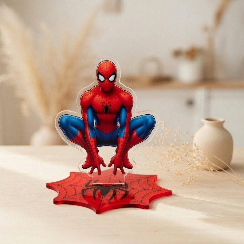 May include: A clear acrylic Spider-Man figure in a crouching pose, with red and blue costume details. The figure is mounted on a red spiderweb base with a black spider emblem. The background is blurred.