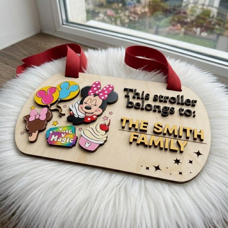 May include: A rectangular light-colored wooden stroller sign with red ribbon loops. It features a Minnie Mouse figure, colorful balloons, a treat, and a ticket labeled Magic. The text reads: This stroller belongs to: THE SMITH FAMILY.