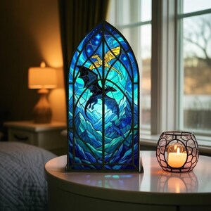 May include: A decorative stained glass window with a dragon design in blue, teal, and gold. The arched window sits on a table next to a candle in a decorative holder. The background includes a lamp and a window overlooking a landscape.