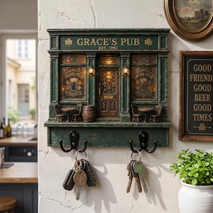Könnte beinhalten: Ein dunkelgrüner Wand-Schlüsselhalter im Design einer irischen Pub-Fassade mit der Aufschrift Grace's Pub Est. 1992. Das Objekt verfügt über zwei kleine Laternen, zwei Tische mit Stühlen, ein Fass und zwei schwarze Metallhaken mit Schlüsseln.