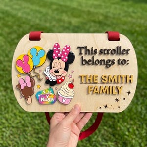 May include: A rectangular wooden stroller sign with rounded corners and red fabric straps. It features a cartoon mouse character, colorful balloons, a cupcake, and a ticket graphic. The text reads: This stroller belongs to: THE SMITH FAMILY.