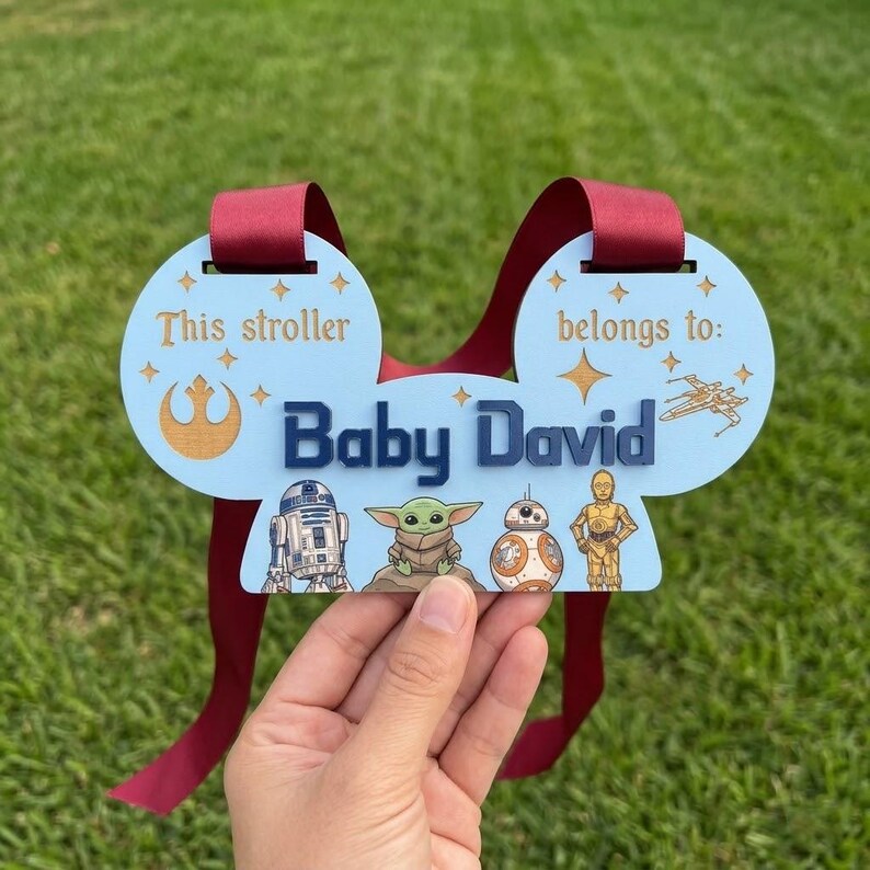 Peut inclure: Une &eacute;tiquette de poussette bleu clair avec le texte "This stroller belongs to: Baby David." Comprend des personnages de Star Wars et un ruban bordeaux pour l'accrocher. L'&eacute;tiquette est tenue sur un fond d'herbe verte.