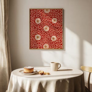 May include: A framed artwork with a red background and a white floral pattern. The artwork is hanging on a white wall. A table is set with a cake, biscuits, and a mug of coffee. The table is covered with a white tablecloth.