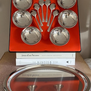 Puede incluir: Un juego de cuencos y cucharas de servir plateados, dispuestos en una caja roja. El juego incluye seis cuencos y seis cucharas. También se ve una bandeja ovalada plateada y libros.