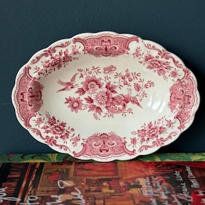 Op de afbeelding: Ovale serveerschaal met een geschulpte rand. De witte keramische schaal heeft een rood bloemen- en vogelontwerp. Het ontwerp omvat bloemen, bladeren en een vogel in vlucht. De rand van de schaal heeft een decoratieve rand.