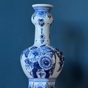 May include: A tall ceramic vase featuring a white base with intricate blue hand-painted floral patterns and a bird illustration. The vase has a bulbous body, a slender neck with a rounded section, and a flared rim, showcasing detailed blue brushwork.