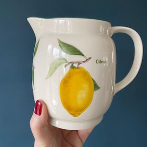 Op de afbeelding: Crèmekleurige keramische kan met een citroenillustratie. De kan heeft een gele citroen met groene bladeren en het woord "Citron". Het heeft een gebogen handvat en een schenktuit.