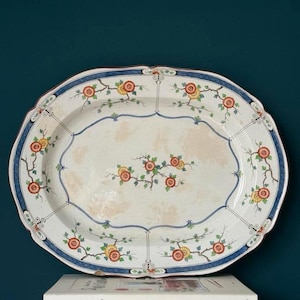May include: An oval, antique serving platter with a white base and a blue and red floral design. The platter has a scalloped edge and is decorated with branches of orange and yellow flowers. The platter is resting on top of two books.
