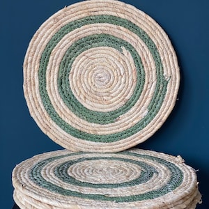 Puede incluir: Un conjunto de salvamanteles redondos y tejidos, de color beige natural con un diseño en espiral verde. Los salvamanteles están hechos de fibra natural y están apilados. Cada salvamanteles mide aproximadamente 30 cm de diámetro.
