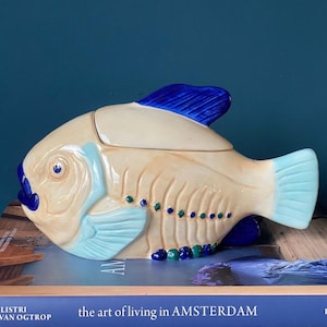 Op de afbeelding: Een keramische visvormige voorraadpot met deksel in een crèmekleur met een glanzende afwerking. De vis heeft een felblauwe rugvin, een lichtblauwe staart en zijvin, en donkerblauwe accenten op de lippen en ogen. Kleine groene en blauwe stippen sieren de zijkant.