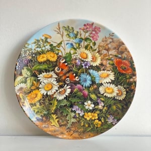 May include: A decorative plate featuring a vibrant floral design with daisies, poppies, and a butterfly. The plate has a light blue border and a detailed painting of a garden scene. The plate is round.