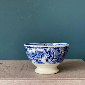 Può includere: Una piccola ciotola in ceramica bianca con un motivo floreale blu e bianco. La ciotola ha una base a piedistallo e si trova su un libro.