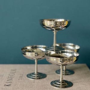 Könnte beinhalten: Vier silberne Champagner-Coupe-Gläser mit reflektierendem Finish sind auf einer neutralen Oberfläche angeordnet. Die Gläser haben ein klassisches Design mit einer breiten, flachen Schale und einem schlanken Stiel. Der Hintergrund ist einfarbig teal.