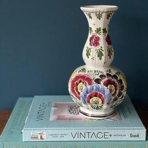 Op de afbeelding: Een crèmekleurige keramische vaas met een geribbelde, bolvormige structuur en handgeschilderde bloemmotieven in rood, blauw, geel en groen. De vaas heeft een smalle hals en een brede voet, versierd met gedetailleerde blad- en bloempatronen.