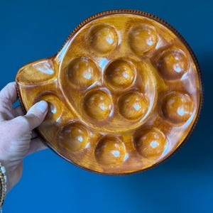May include: A brown ceramic escargot plate with 12 circular indentations, arranged in a spiral pattern. The plate has a handle and a glossy finish. The plate is held against a dark blue background.