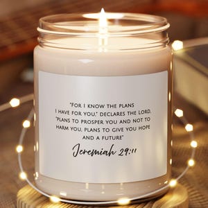 Puede incluir: Una vela encendida en un frasco de vidrio transparente con una etiqueta blanca. La etiqueta tiene texto de Jeremías 29:11. La vela está sobre un posavasos de madera pequeño y redondo. Una cadena de luces y una guitarra están en el fondo.