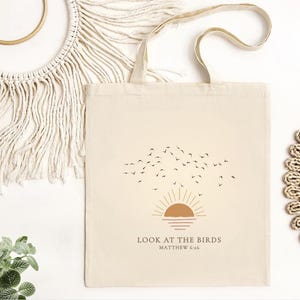 May include: Cream-colored tote bag featuring a sunset design with birds in flight. The text "LOOK AT THE BIRDS" and "MATTHEW 6:26" are printed below the graphic. The bag has a simple, minimalist aesthetic.
