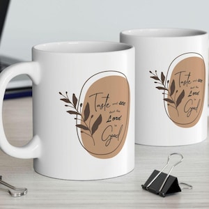 May include: Two white ceramic mugs with a brown oval design and the text "Taste and see that the Lord is Good". The design includes brown leaves and stems. The mugs are on a light-colored surface.