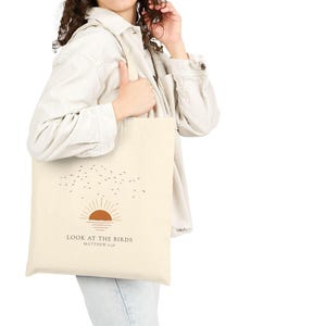 May include: Cream-colored tote bag featuring a minimalist design with a brown sun, birds, and the text "LOOK AT THE BIRDS" and "MATTHEW 6:26". The bag has a simple, rectangular shape and long straps.