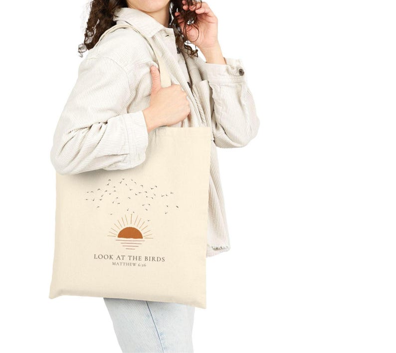 May include: Cream-colored tote bag featuring a minimalist design with a brown sun, birds, and the text "LOOK AT THE BIRDS" and "MATTHEW 6:26". The bag has a simple, rectangular shape and long straps.