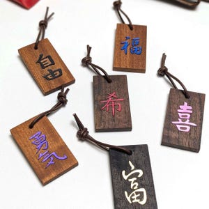 Op de afbeelding: Zes rechthoekige houten labels met donkerbruine leren koorden. Elk label heeft een uniek gegraveerd Chinees karakter gevuld met gekleurde inleg: zwart, blauw, rood, paars, roze en goud. De houtnerf is op elk stuk duidelijk zichtbaar.