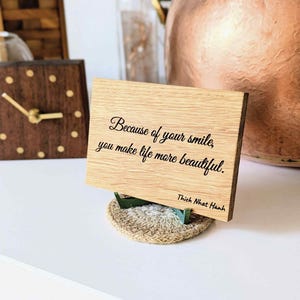Könnte beinhalten: Eine rechteckige, helle Holztafel mit dem schwarz aufgedruckten Text "Because of your smile, you make life more beautiful." und "Thich Nhat Hanh." Die Tafel wird von einem kleinen, runden, strukturierten, beigen geflochtenen Sockel gestützt.