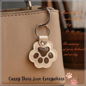 Könnte beinhalten: Ein beigefarbener Pfoten-Schlüsselanhänger mit einem silbernen Ring, der an einer beigefarbenen Ledertasche befestigt ist. Der Schlüsselanhänger hat eine transparente Mitte und genähte Kanten. Der Text "Carry Their Love Everywhere" steht unten.