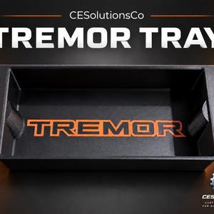 May include: A rectangular black tray with the word TREMOR printed in orange on the interior base. The tray features a wide rim and a deep storage compartment. The text CESolutionsCo and TREMOR TRAY appear in white above the tray.