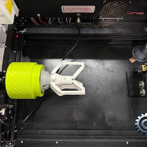 May include: A close-up of a 3D printed robotic arm with a yellow gripper wheel. The arm is attached to a black and silver machine with a black and white gear logo. The text "Turn off device before plugging/unplugging" is visible on the machine.