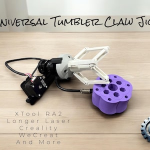 May include: A white robotic claw arm with a purple, multi-hole jig. The arm is connected to a black motor and wires. The text "UNIVERSAL TUMBLER CLAW JIG" is at the top of the image. The text "XTool RA2 Longer Laser Creality WeCreat And More" is at the bottom of the image.