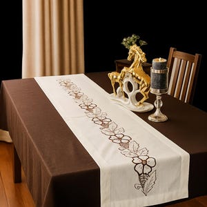 May include: A brown tablecloth with a white table runner featuring embroidered floral designs. A gold horse figurine and a candle holder with a lit candle are on the table. A wooden chair is visible in the background.