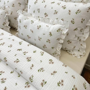 May include: White floral bedding set featuring a duvet cover and pillows with frilled edges. The fabric has a textured, waffle-like design, and the floral pattern includes small, brown and green flowers. The set is designed for a bed.