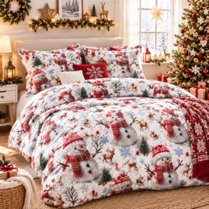 May include: A white bedding set featuring a repeating pattern of snowmen wearing red knit scarves and hats, interspersed with small brown deer, bare trees, and blue snowflakes. A solid red accent pillow with a white snowflake design is centered.
