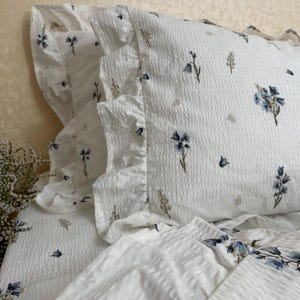 May include: White bedding set with a floral pattern in blue and tan. The set includes a pillowcase with a ruffled edge and a textured surface. The floral design is scattered across the fabric.