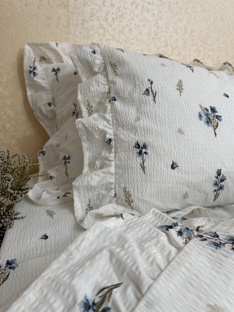May include: White bedding set with a floral pattern in blue and tan. The set includes a pillowcase with a ruffled edge and a textured surface. The floral design is scattered across the fabric.