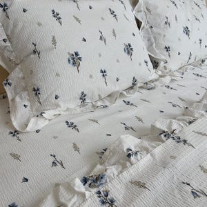 May include: White bedding set with a floral pattern in blue and gray. The set includes two pillowcases and a fitted sheet, all with a ruffled edge. The fabric has a textured, waffle-like appearance.