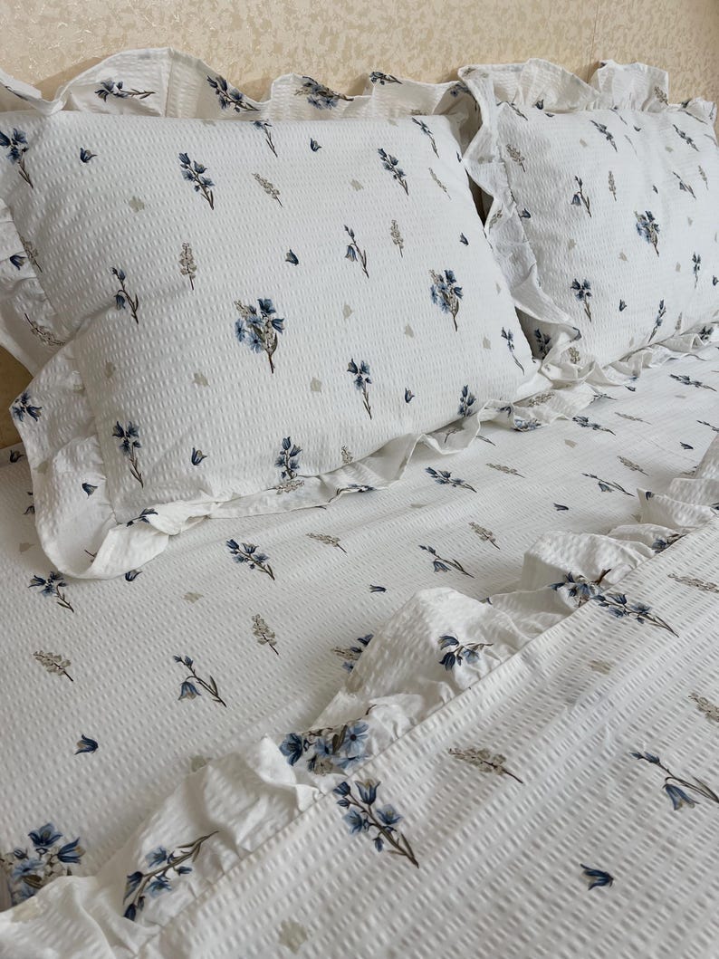 May include: White bedding set with a floral pattern in blue and gray. The set includes two pillowcases and a fitted sheet, all with a ruffled edge. The fabric has a textured, waffle-like appearance.