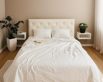 May include: A neatly made bed with white bedding, including a duvet cover and two pillows. The headboard is cream-colored with button tufting. A small book rests on the bed. Two floating white nightstands flank the bed, each with a plant.
