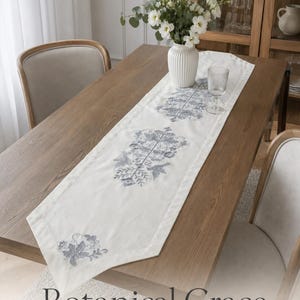 May include: A cream-colored table runner measuring 160x38 cm, featuring intricate blue-grey botanical and bird embroidery patterns. The runner is laid flat on a wooden table, showcasing symmetrical floral motifs and pointed ends with decorative stitching.