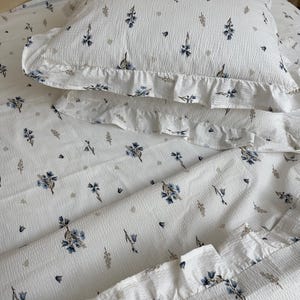 May include: White bedding set with a floral pattern. The set includes a pillowcase and a sheet, both featuring a repeating design of blue flowers and small gray accents. The pillowcase has a ruffled edge. The fabric has a textured appearance.