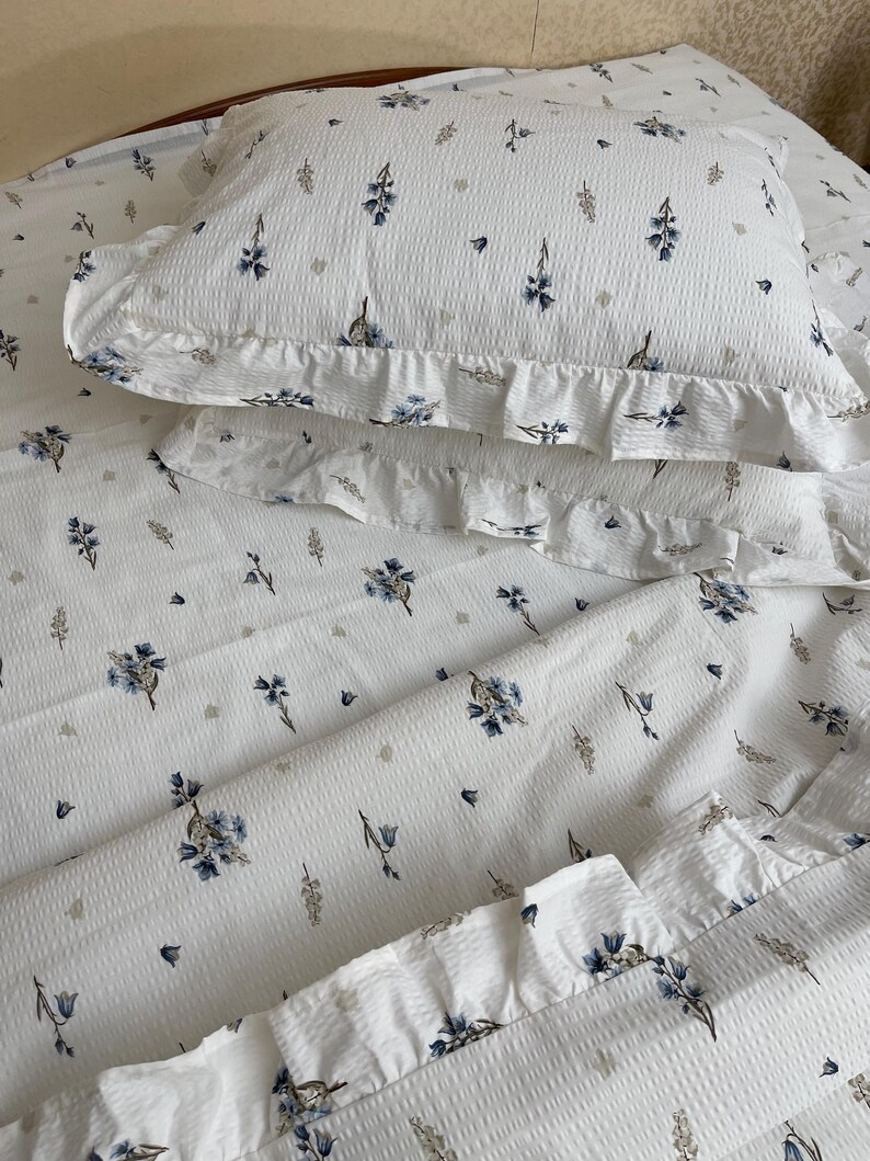 May include: White bedding set with a floral pattern. The set includes a pillowcase and a sheet, both featuring a repeating design of blue flowers and small gray accents. The pillowcase has a ruffled edge. The fabric has a textured appearance.