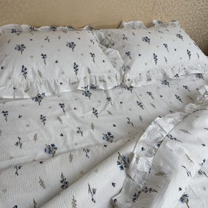 May include: White bedding set with a floral pattern featuring blue and gray flowers and foliage. The set includes two ruffled pillowcases and a duvet cover. The fabric has a textured appearance.