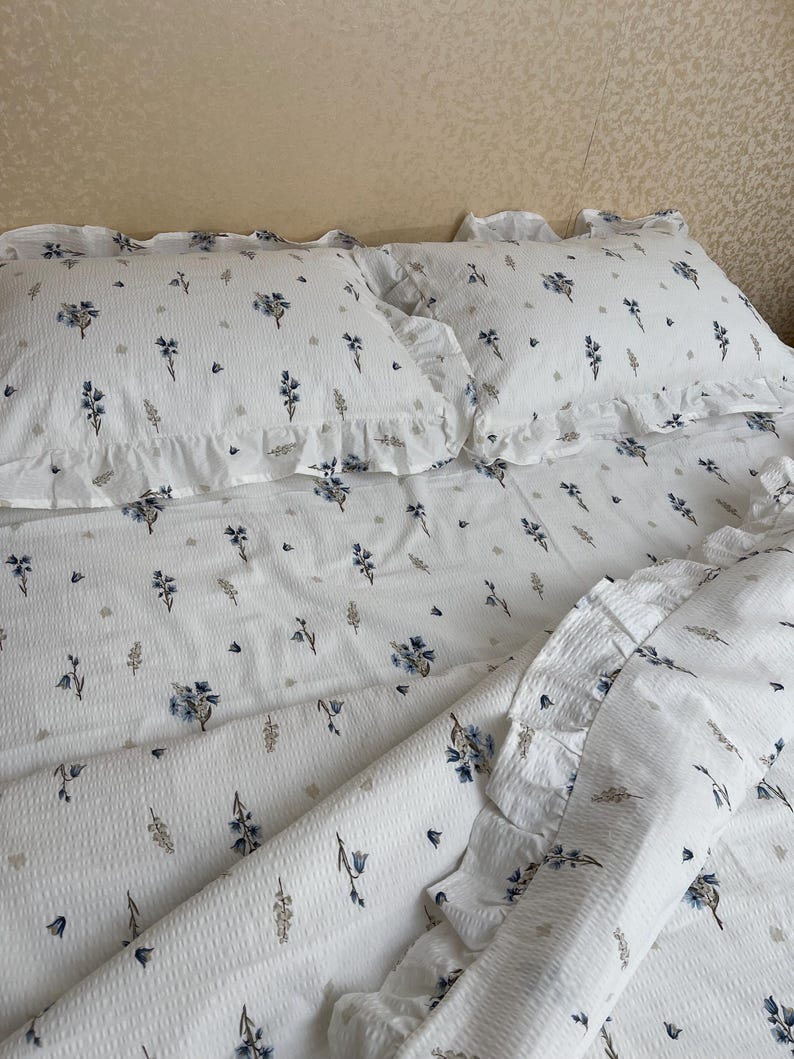 May include: White bedding set with a floral pattern featuring blue and gray flowers and foliage. The set includes two ruffled pillowcases and a duvet cover. The fabric has a textured appearance.